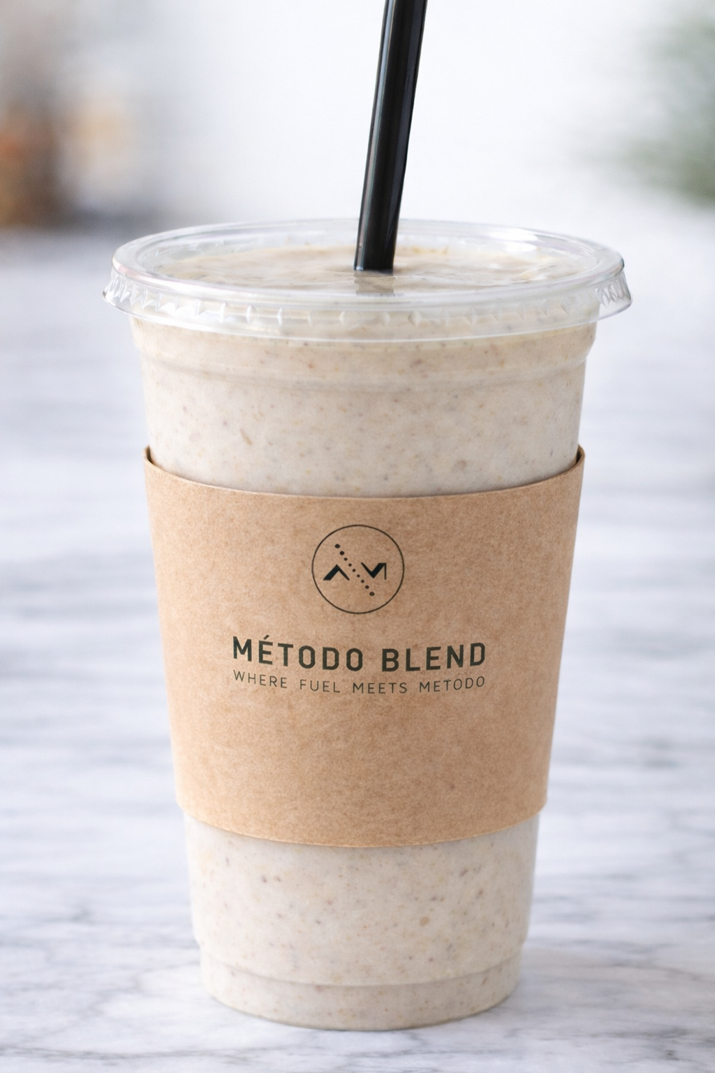 A cold beverage in a clear plastic cup with a black straw, wrapped in a brown paper sleeve with the logo 'Método Blend' and the tagline 'Where fuel meets metodo,' placed on a white textured surface.