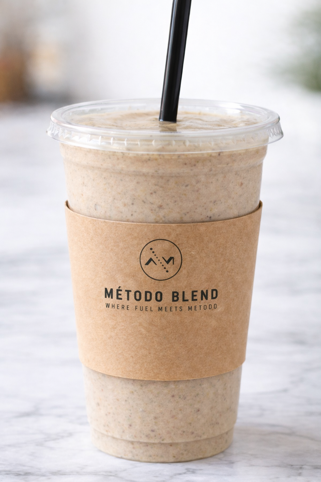 A blended coffee drink in a clear plastic cup with a black straw, wrapped in a brown paper sleeve with a logo and the words 'MÉTODO BLEND' and 'WHERE FUEL MEETS METODO'.
