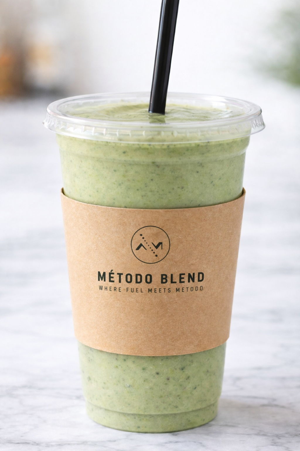 A green smoothie in a clear plastic cup with a black straw, wrapped with a brown paper sleeve labeled 'MÉTODO BLEND' and the subtitle 'WHERE FUEL MEETS METODO,' sitting on a marble surface.