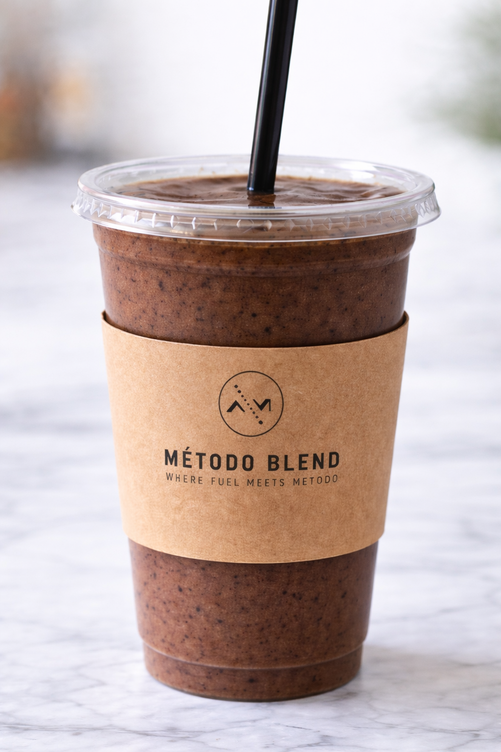 A plastic cup with a black straw filled with an iced chocolate drink, wrapped with a brown paper sleeve that has the Métdo Blend logo and slogan.