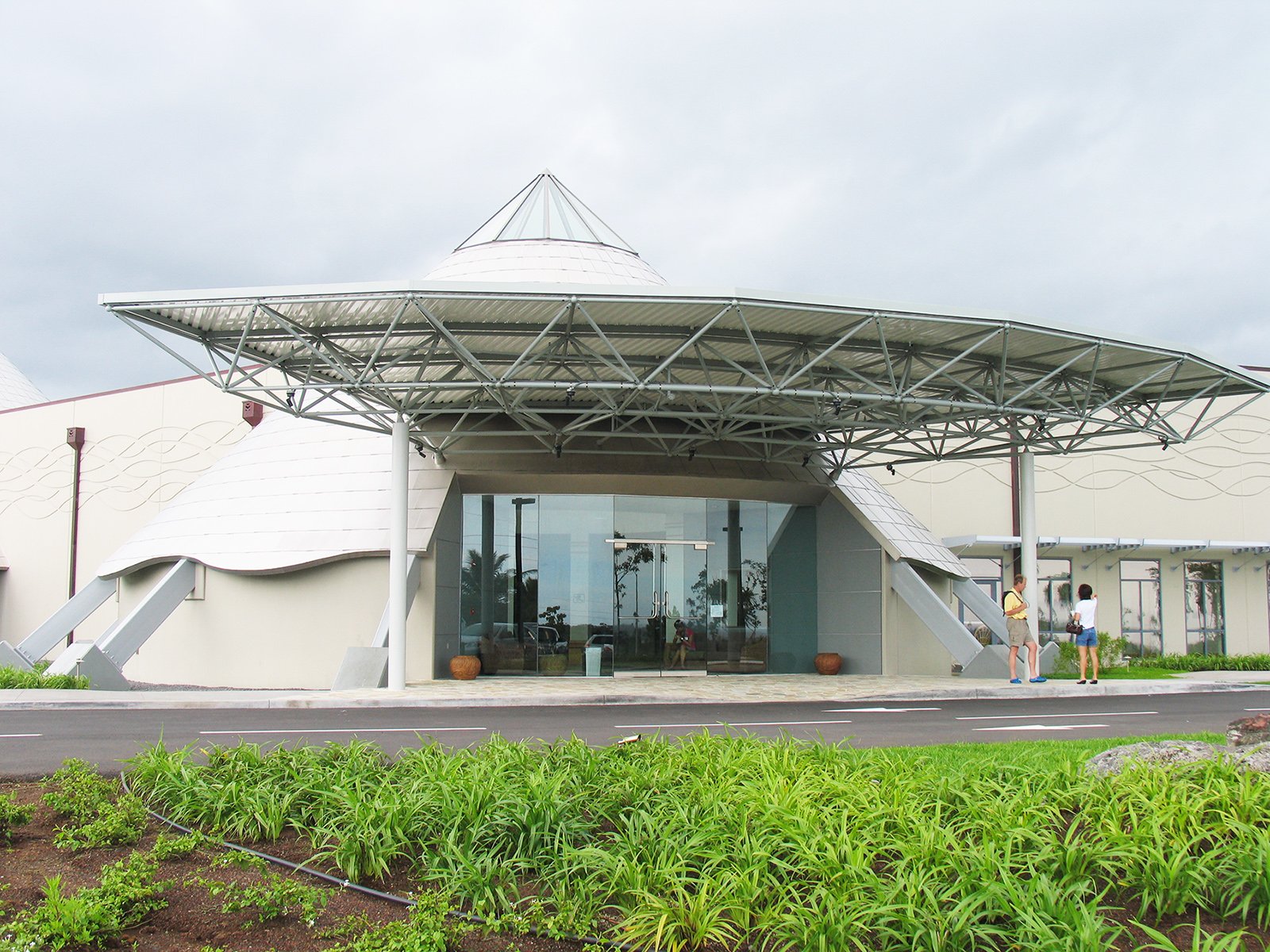 Modern building with a unique architectural roof structure and large glass entrance, surrounded by greenery and a few people standing outside.