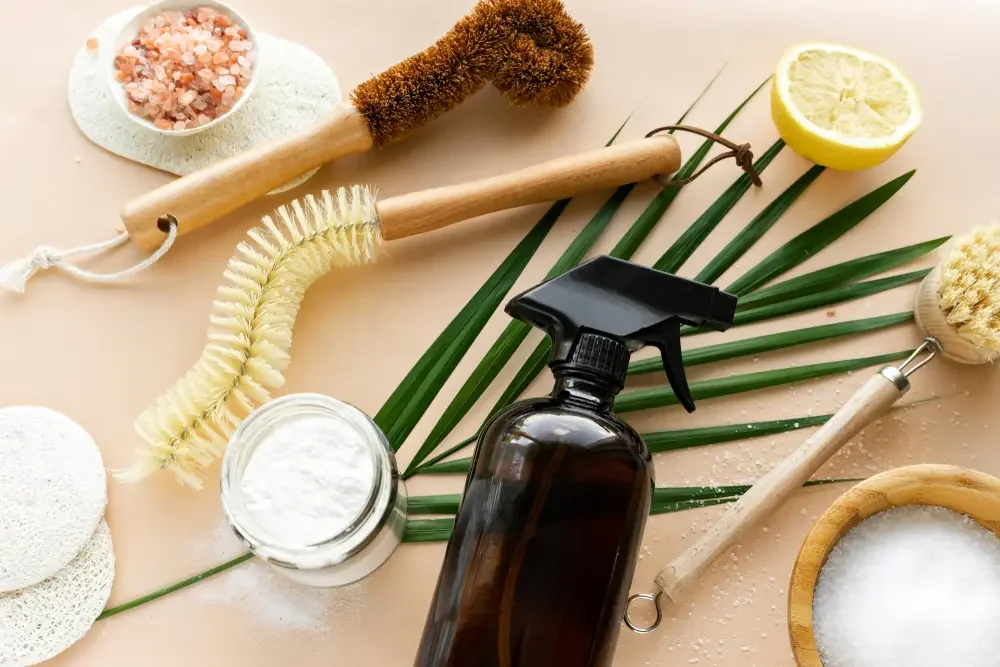 Natural cleaning supplies including a spray bottle, lemon half, brushes, sponges, salt, and cleaning cloths on a light background.