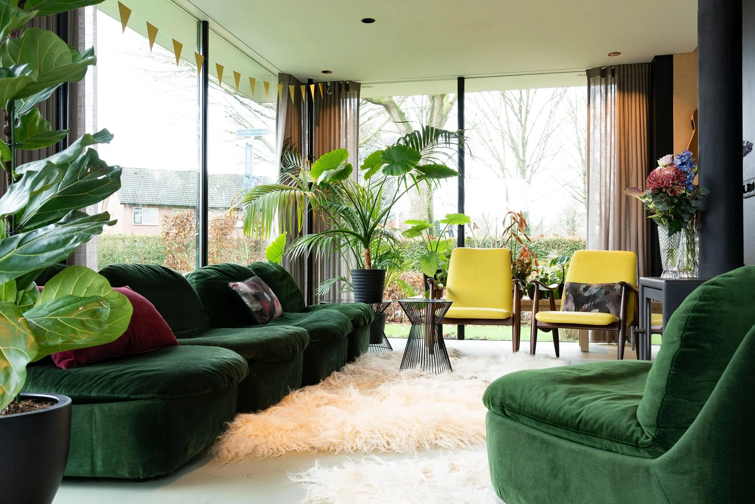 Living room with large windows, green velvet sofa, yellow armchairs, potted plants, floral arrangement, shaggy white rug, and natural light.