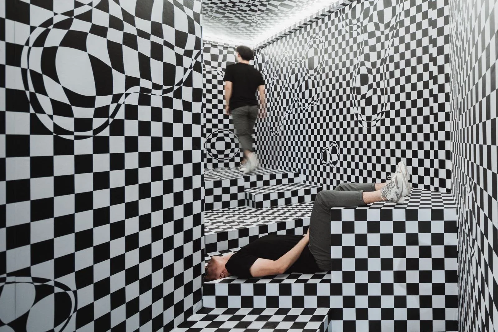 An optical illusion room with black-and-white checkered walls, floors, and stairs. A person is lying down on a platform, and another person is walking away on the stairs.