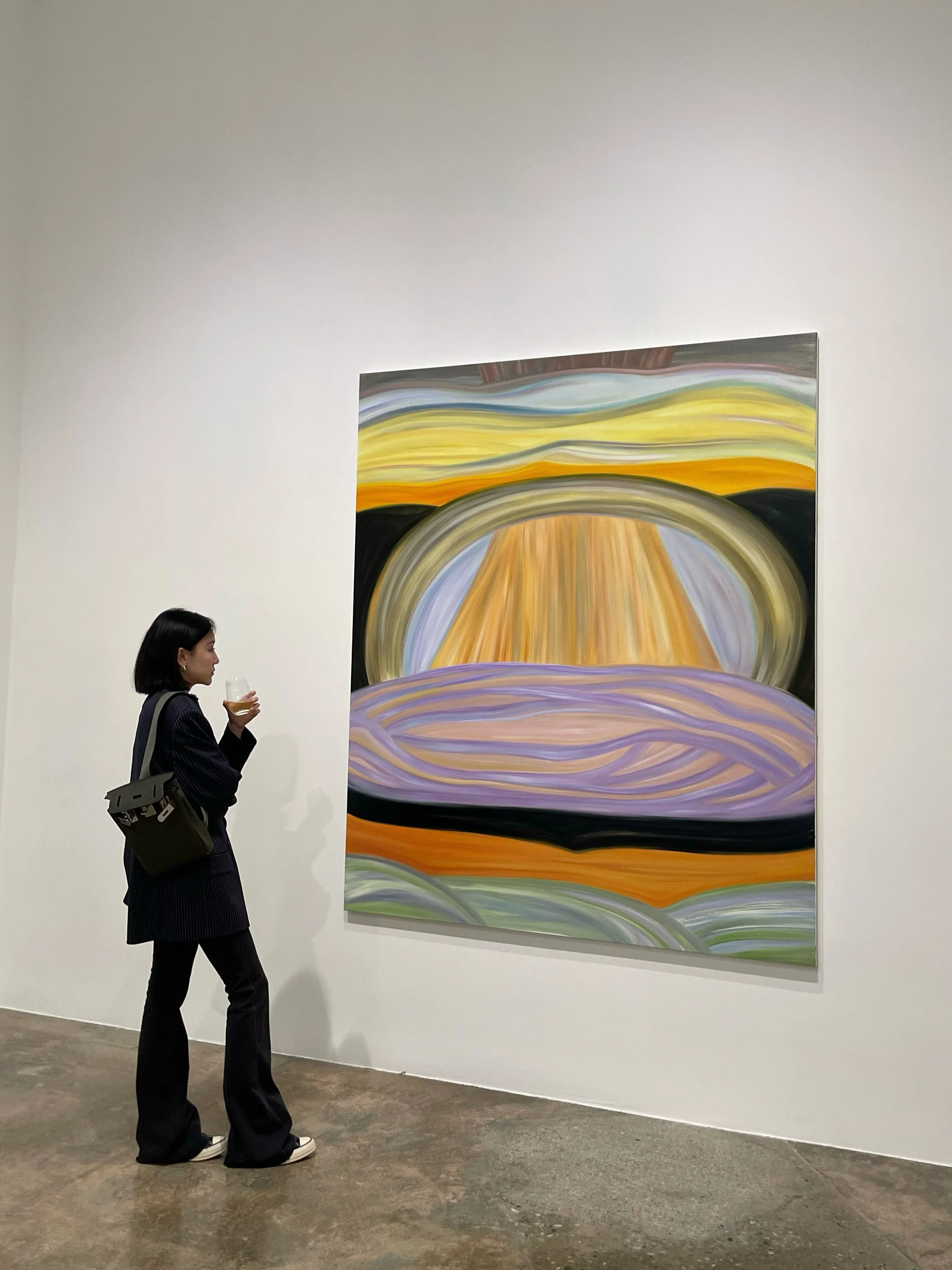A woman with short black hair, dressed in a black pinstripe suit and white shoes, is standing in an art gallery holding a drink, looking at an abstract painting with swirling colors and patterns on a white wall.