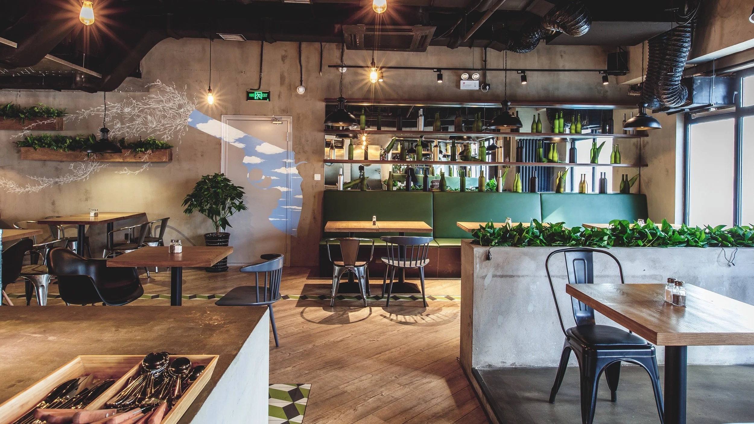 Interior of a modern cafe with wooden tables, metal chairs, green seating bench, decorative shelves with bottles and books, potted plants, and large window with natural light.