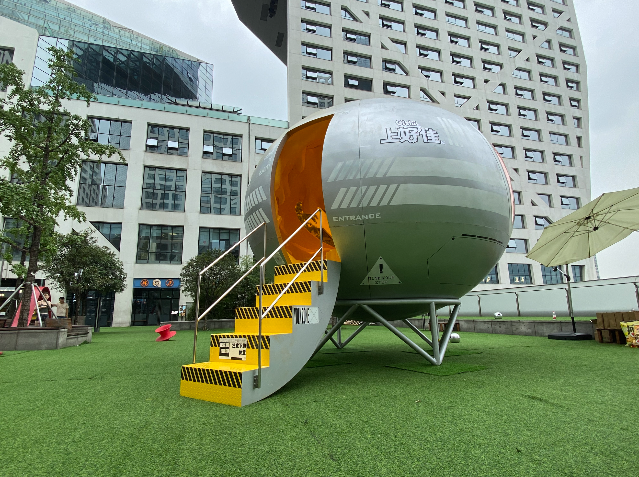 Futuristic spherical structure with stairs leading to an entrance, situated on a grassy area in an urban environment.