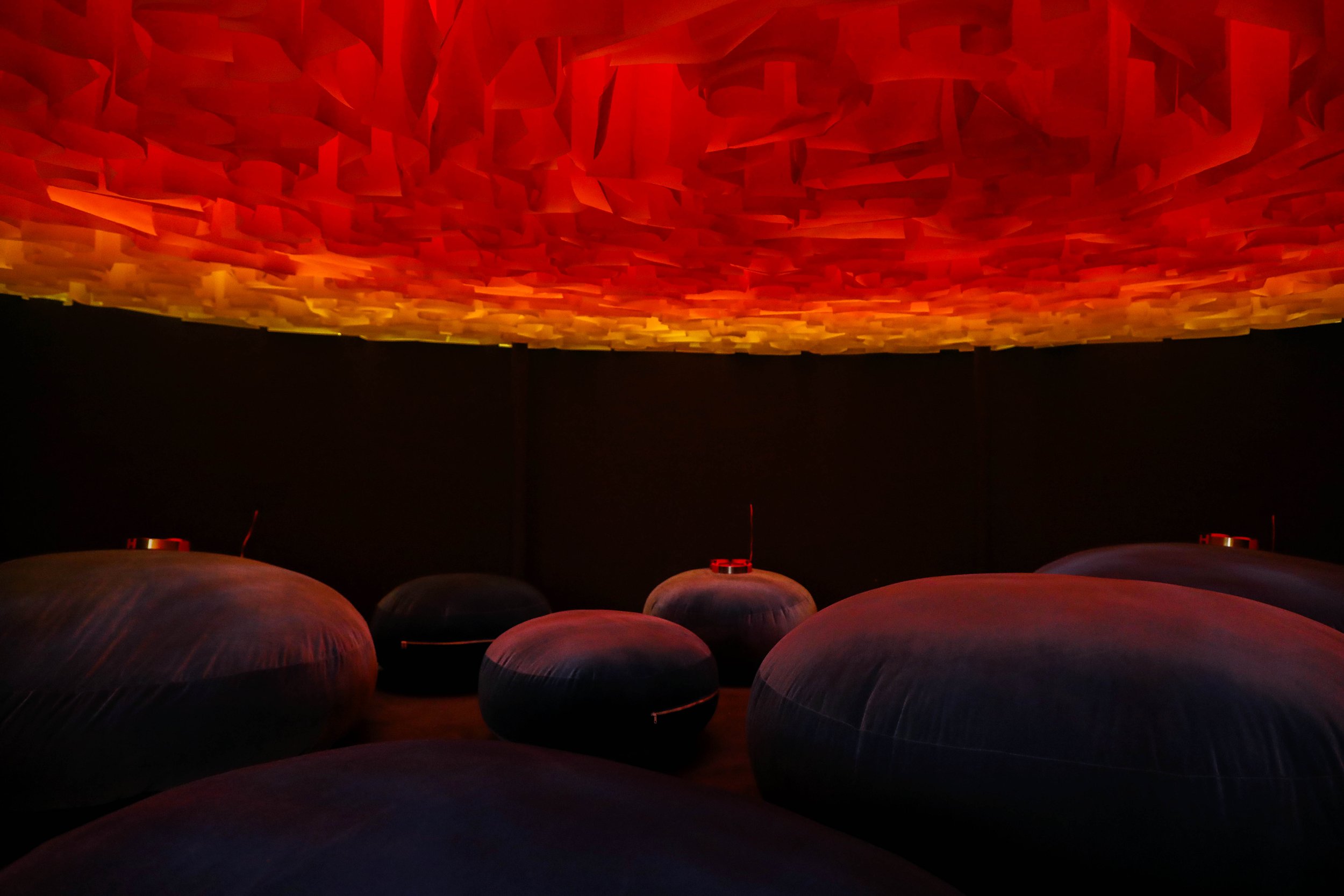 An interior of a dark room with soft LED lighting, featuring several large, rounded, pillow-like bean bags and a suspended ceiling panel emitting a warm orange and red glow.