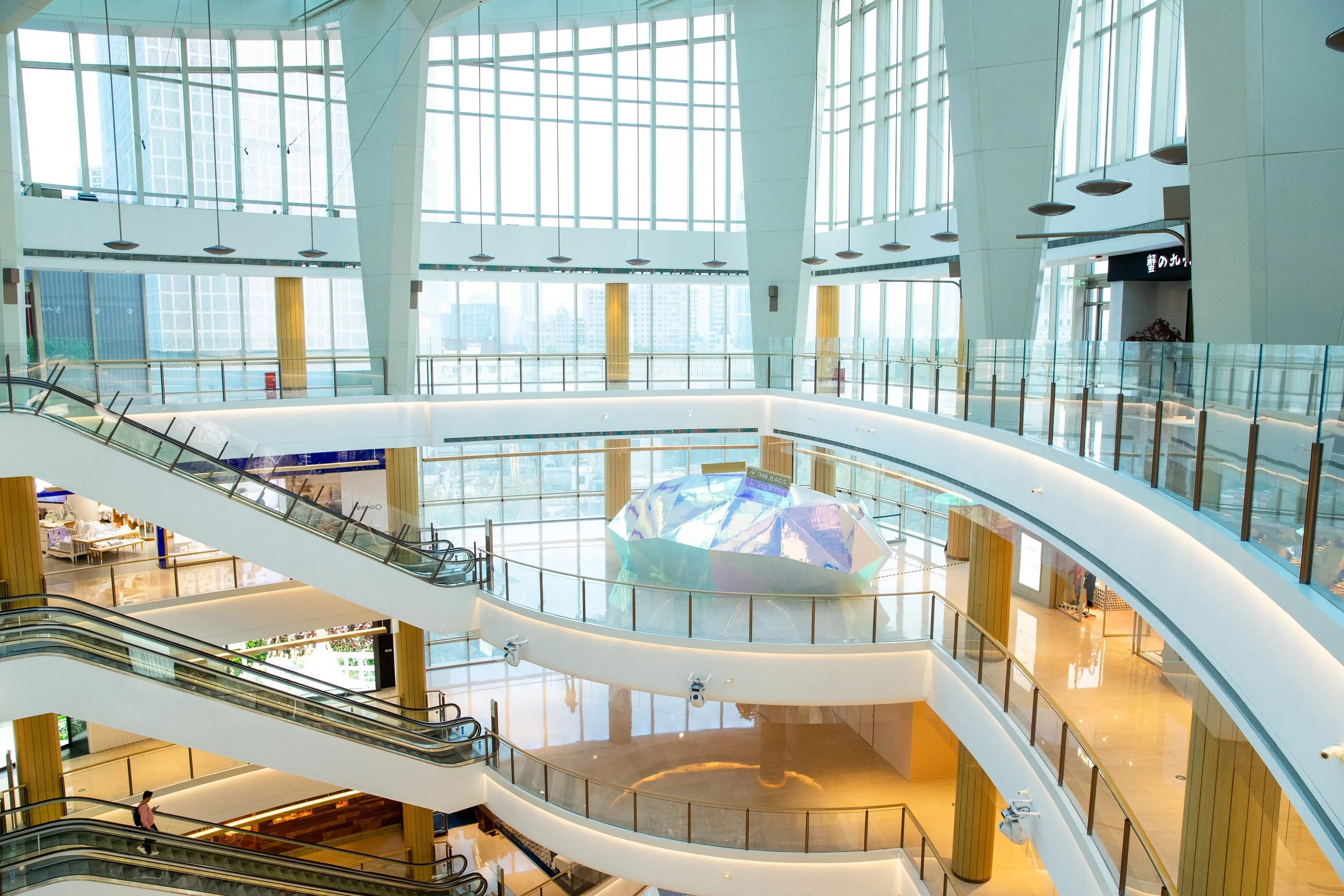 Interior of a modern shopping mall with multiple floors, large glass windows, escalators, and a holographic art installation in the center.