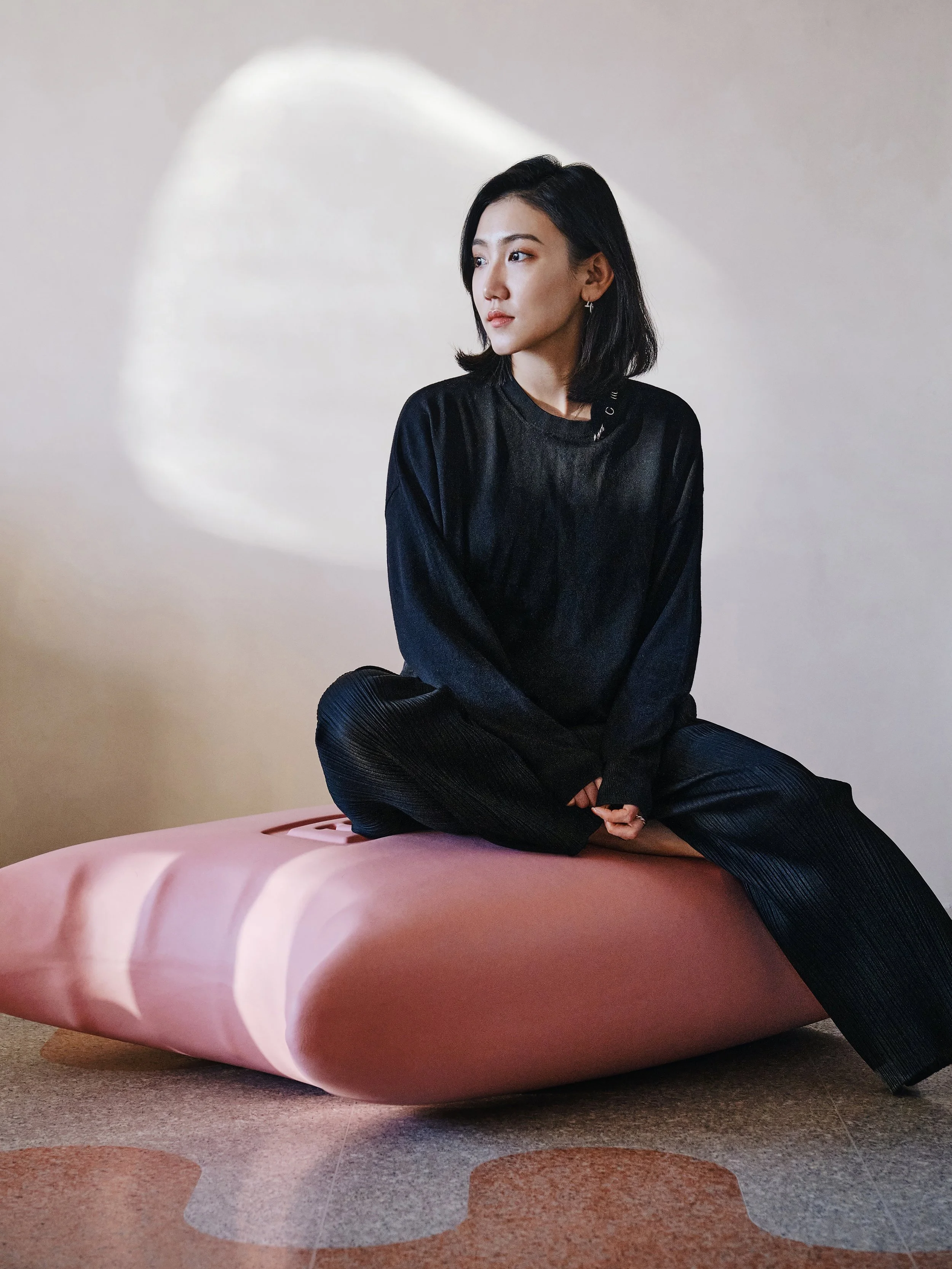 A young woman with black hair sits cross-legged on a pink sculpture that resembles a giant capsule or pill. She is wearing a black satin-like top and black pleated pants, looking to the side with a neutral expression. A circular light spot is projected on the wall behind her.