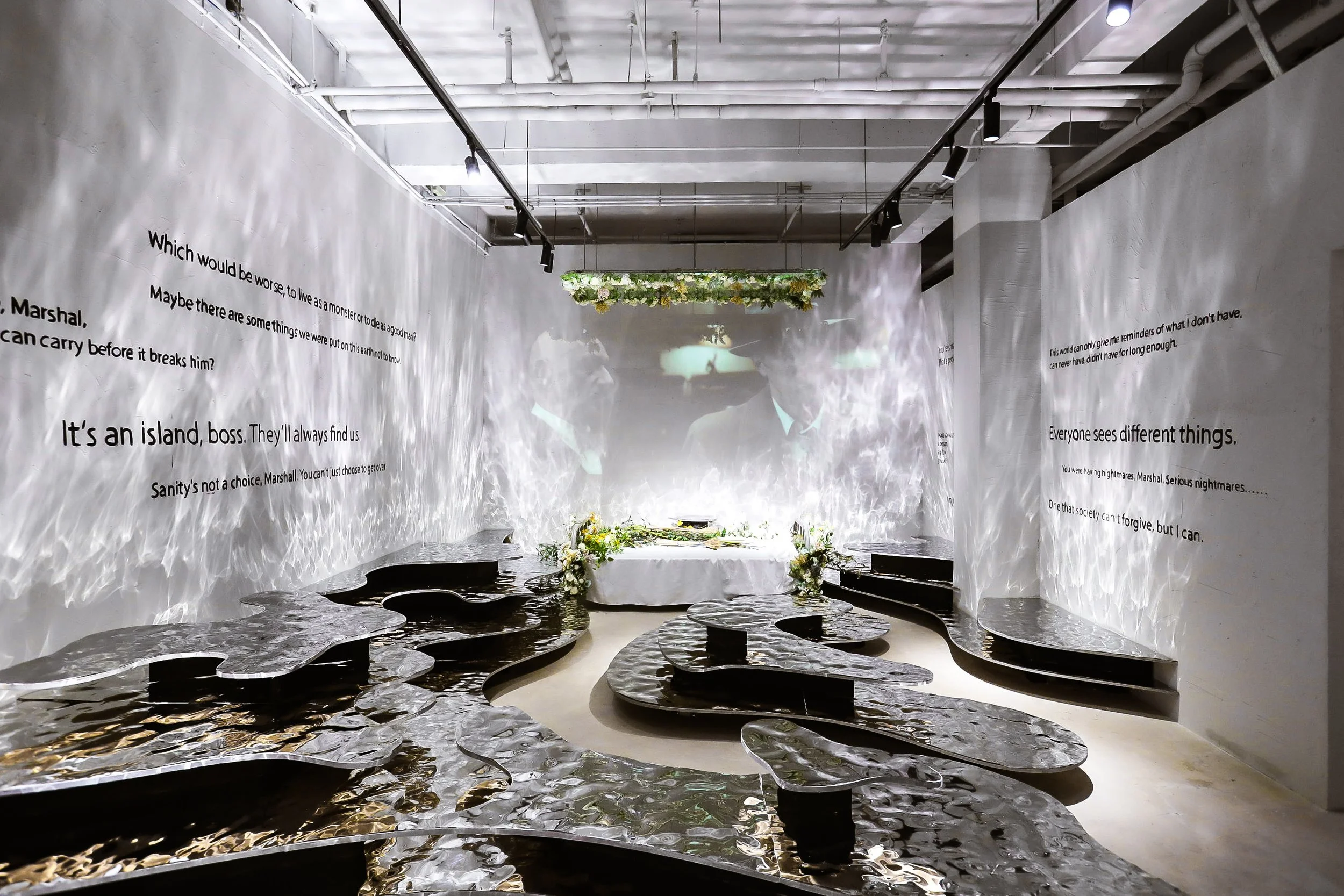 An art installation with reflective, wavy metal floors, white walls with black text, a table with floral arrangements, and a projected video on the wall with hanging greenery above.
