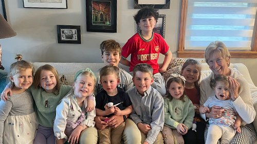 A woman with multiple children sitting on a couch and one standing behind, smiling in a living room with wall art and windows.