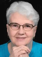 A senior woman with short white hair and glasses, wearing a blue top, smiling gently, with her hand touching her chin.