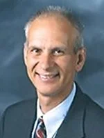 A smiling middle-aged man with gray hair, wearing a suit and tie, standing against a blue background.
