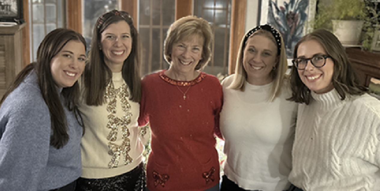 A group of five women standing closely together indoors, smiling at the camera.