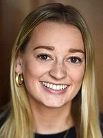 A young woman with blonde hair smiling, wearing gold hoop earrings, against a neutral background.