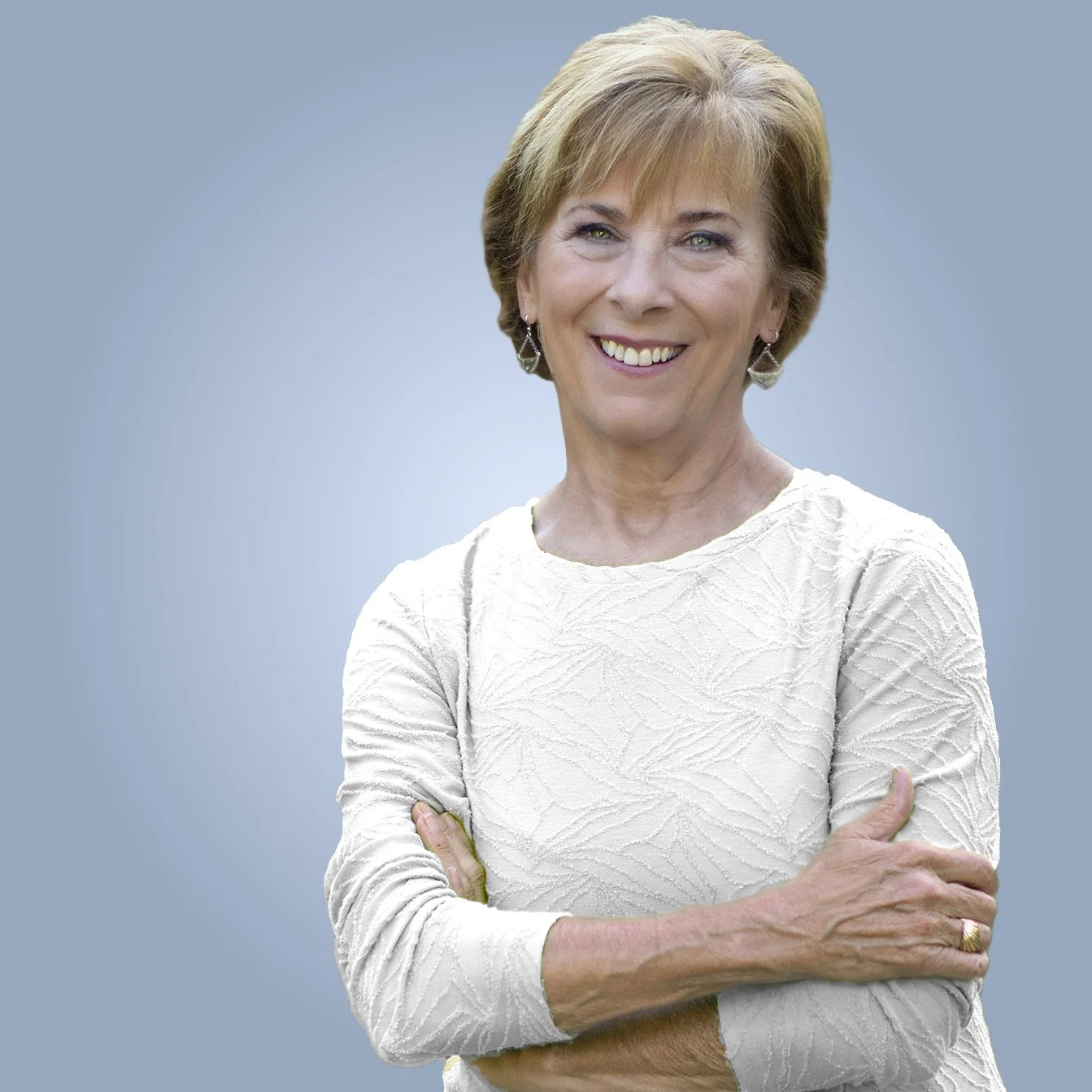 Smiling woman with short blonde hair wearing a white textured top and earrings, crossing her arms in front of a blue-gray background.