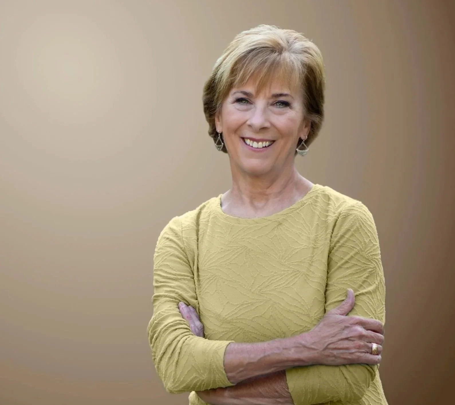 A smiling woman with short, light brown hair wearing a yellow textured long-sleeve dress, posing with arms crossed against a plain beige background.