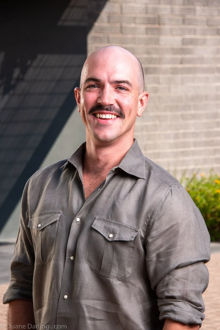 A smiling man with a bald head, mustache, wearing a gray button-up shirt with rolled-up sleeves, standing outdoors in front of a brick wall and greenery.