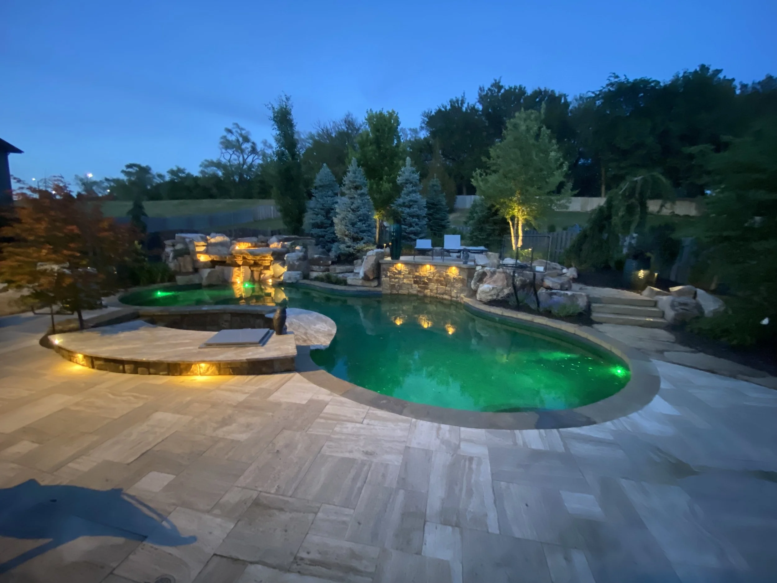 Nighttime view of a backyard swimming pool with built-in lighting, surrounded by stone and tile patio, with trees and landscape in the background.