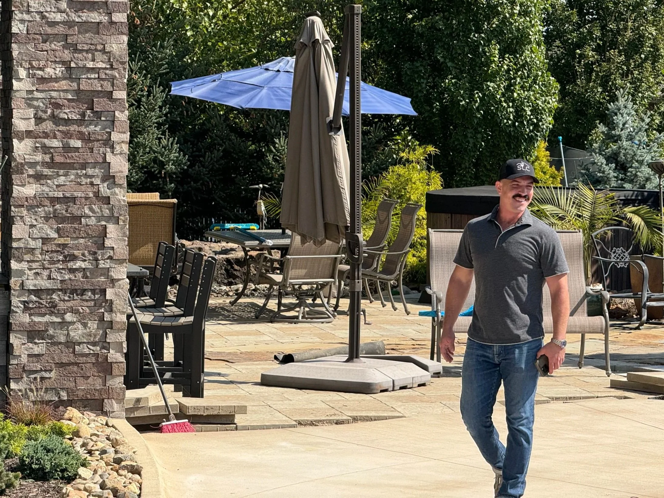 A man in a gray polo shirt, jeans, and a black cap is smiling while walking in a backyard patio. The patio has outdoor furniture, a large umbrella, trees, and lush greenery.