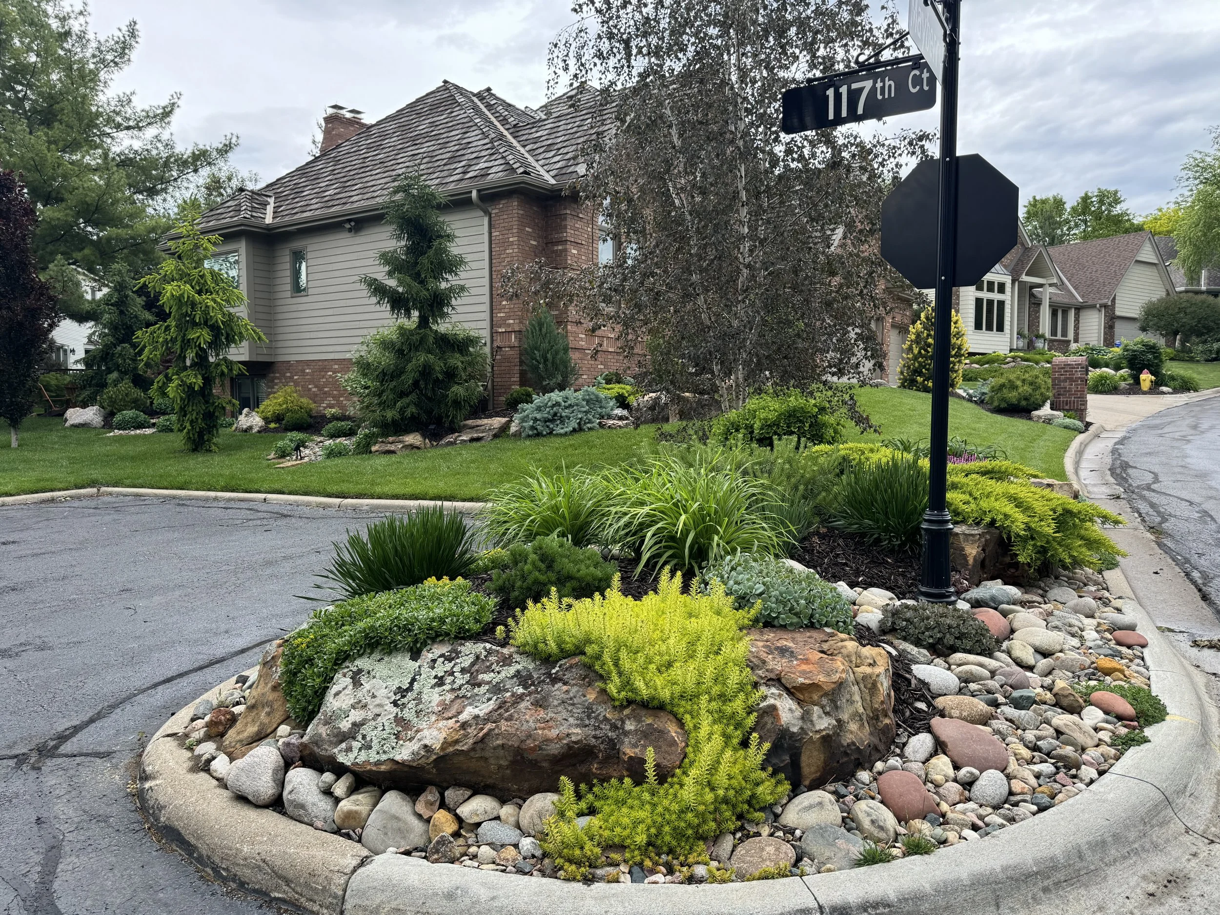 A residential street corner with a landscaped traffic island containing various plants and rocks, a street sign for 117th Ct, and houses with well-maintained yards in the background under an overcast sky.