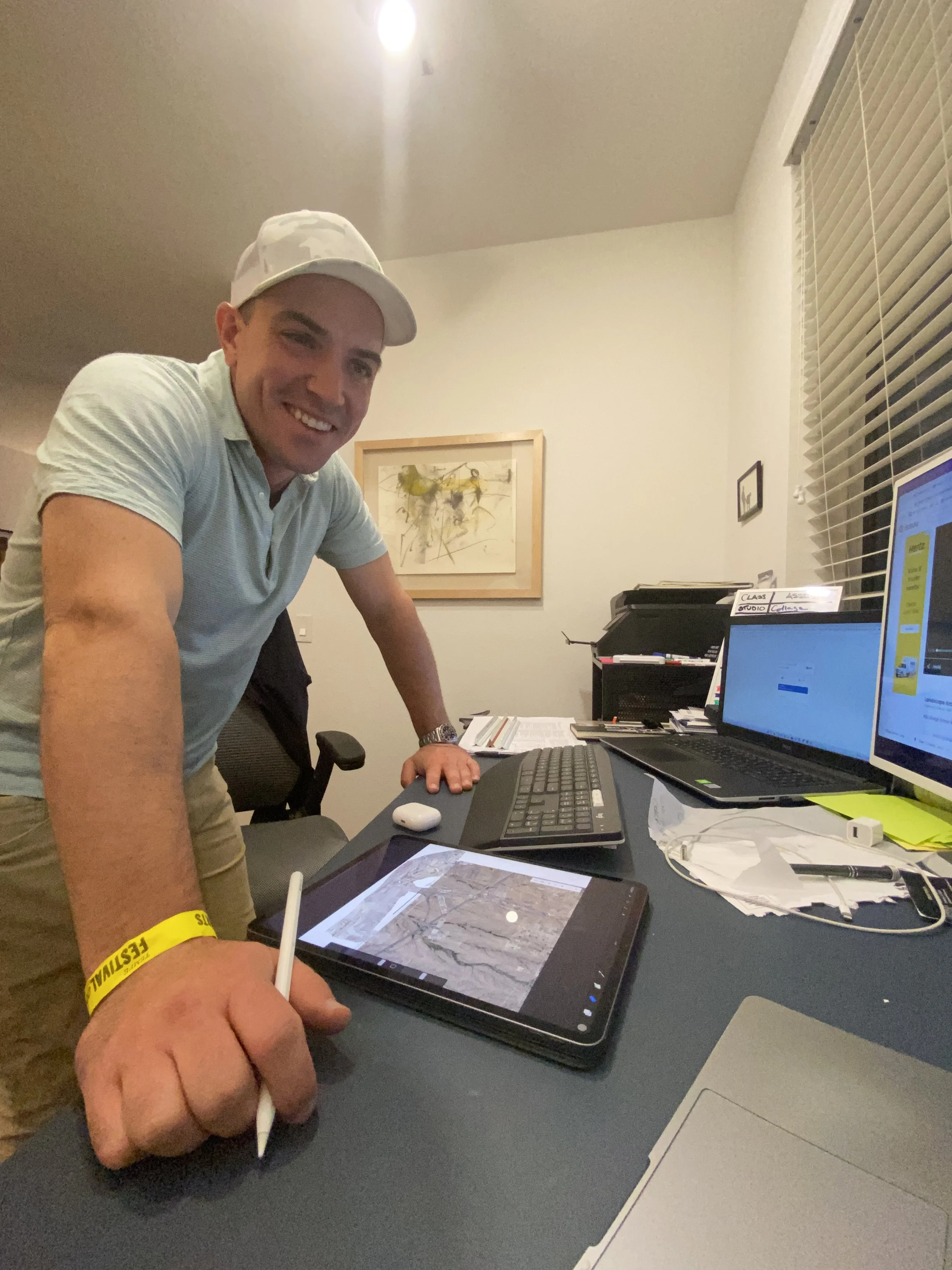 Man in casual attire leaning over a desk, smiling, with a tablet and stylus, surrounded by computer monitors, papers, and office supplies in a home office.
