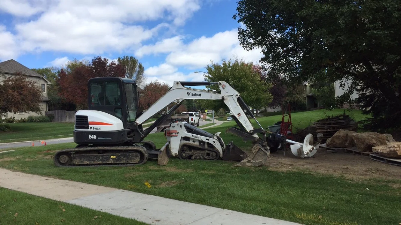 A Bobcat E45 mini excavator with a white attachment digging into a grassy yard in a suburban neighborhood.