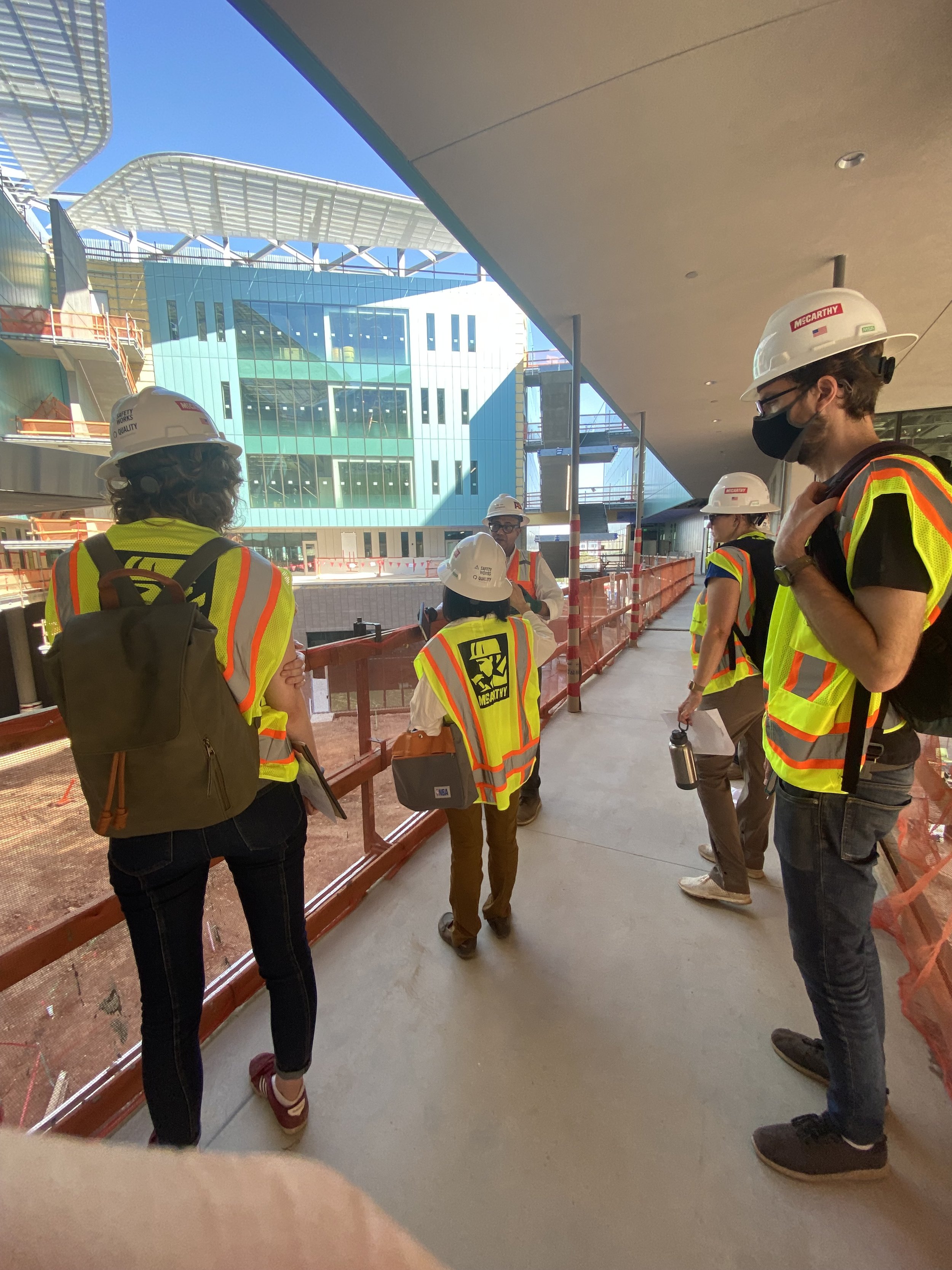Group of construction workers and engineers wearing safety vests, helmets, and masks on a construction site with a modern building in the background.