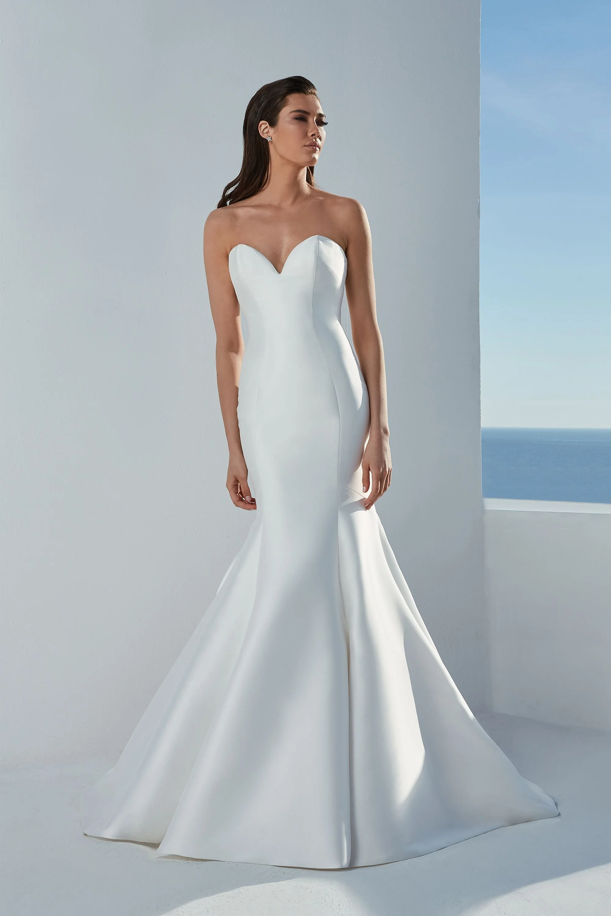 Mujer con vestido de novia blanco de tirantes cortos y escote corazón, de pie en un fondo de paredes blancas y cielo azul, con vista al mar.