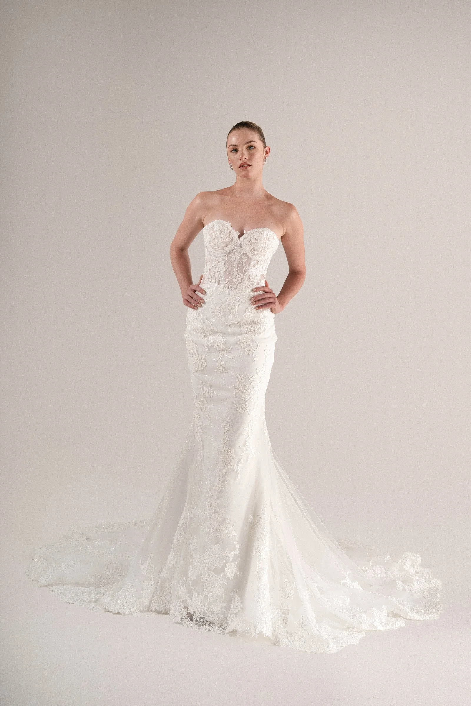 Mujer con vestido de novia elegante y ajustado, con encaje y tren que se extiende en el suelo, posando con las manos en las caderas, en fondo neutro.