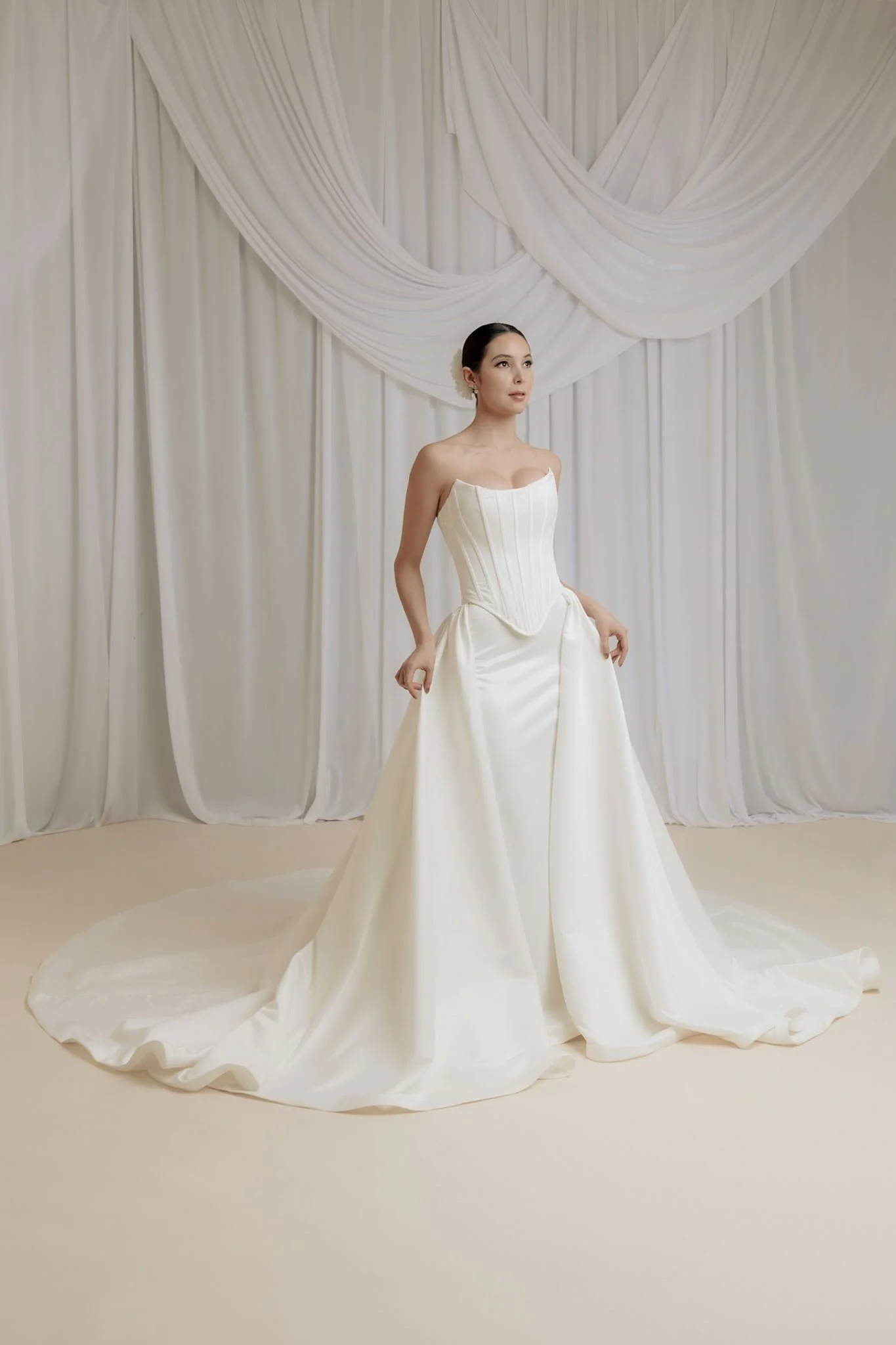 Mujer con vestido de novia, de pie frente a cortinas blancas, en una sesión de fotos de boda.