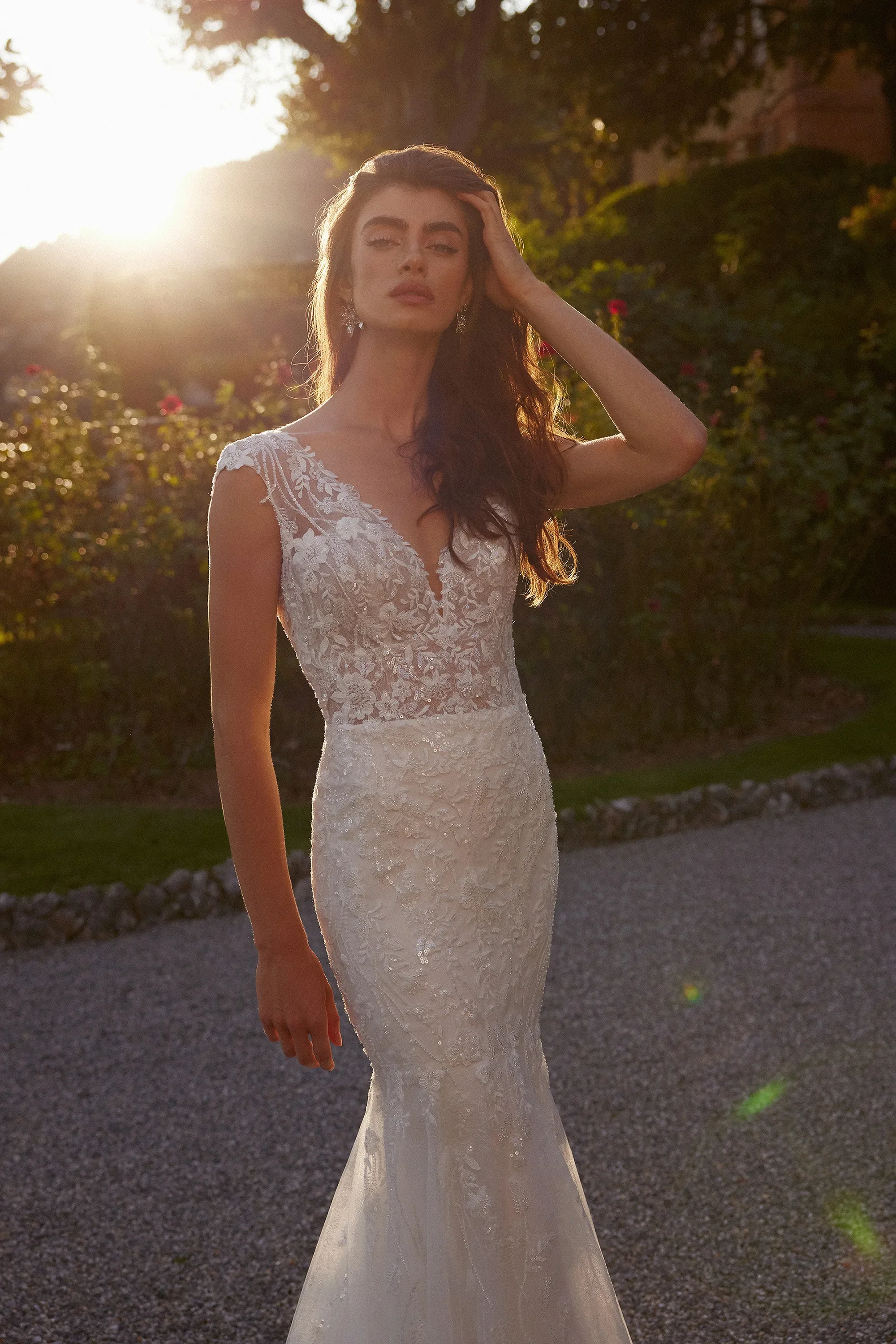 Mujer con vestido de novia largo y encaje, de cabello castaño, posando al atardecer en un jardín con árboles y flores.