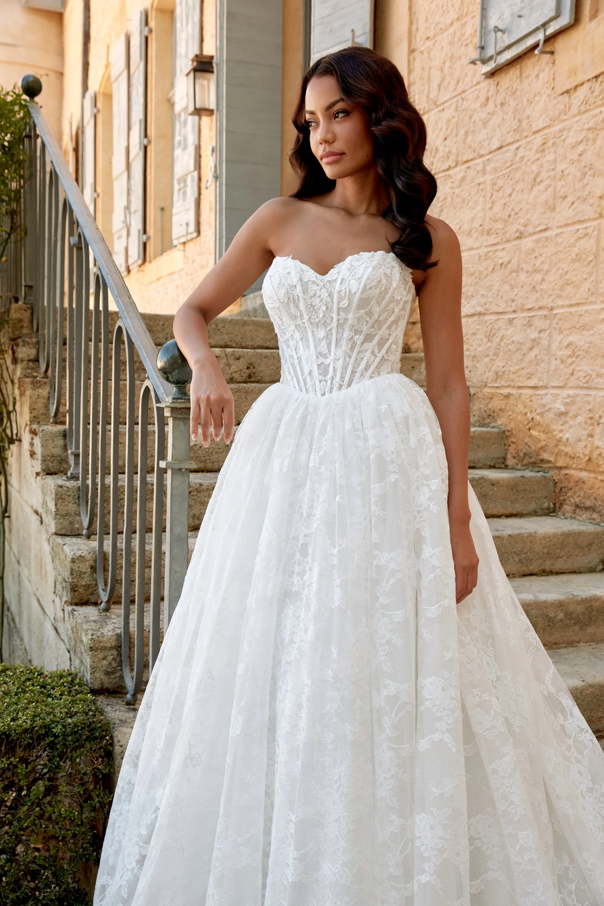Mujer con vestido de novia de encaje en un exterior con escaleras y paredes de ladrillo beige.