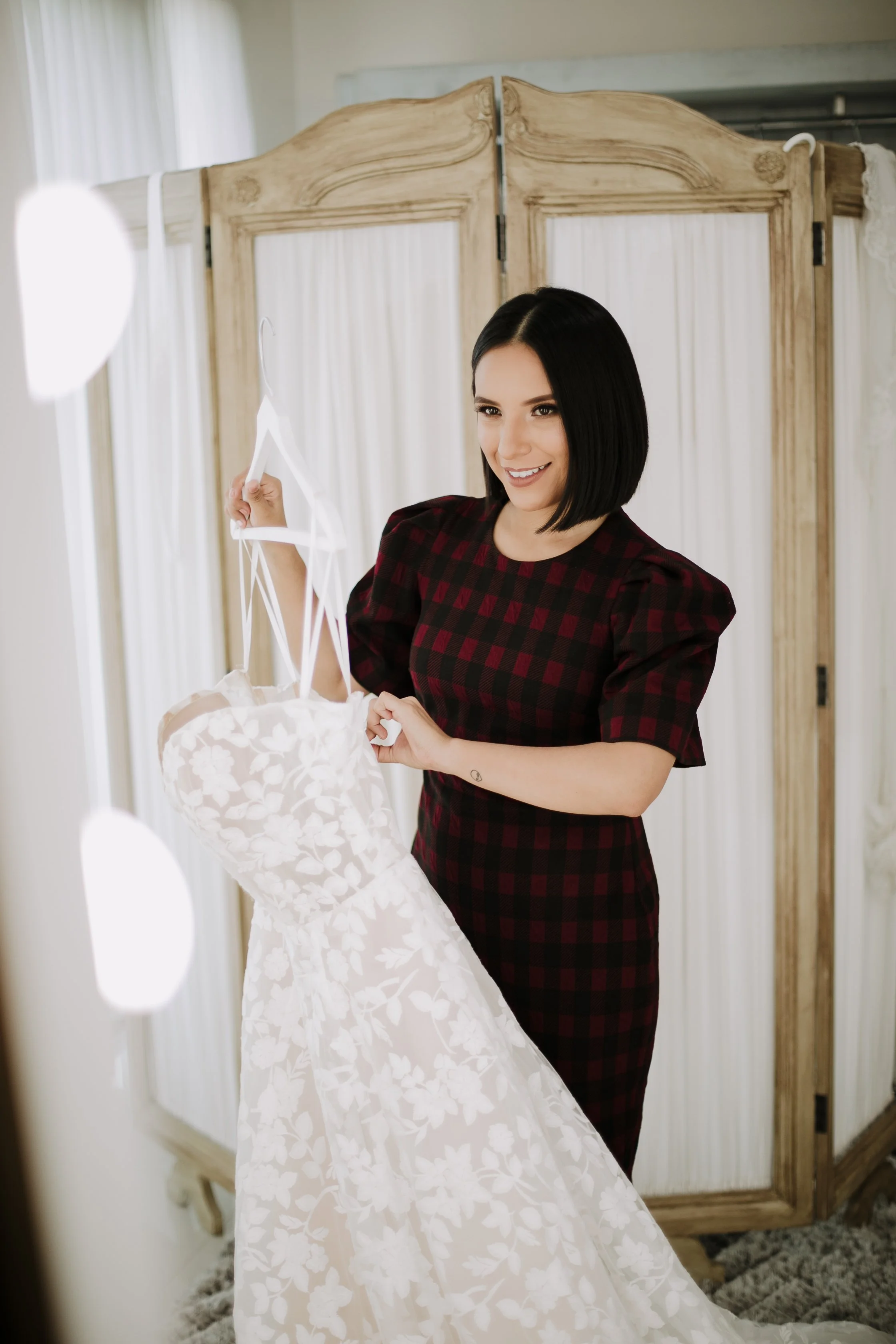 Laura Berino, sonriendo, usando vestido de cuadros rojos y negros, sosteniendo un vestido de novia blanco con encaje floral, en un vestidor con separador de madera.