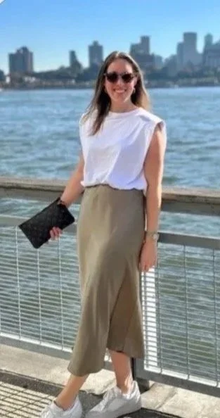A woman wearing sunglasses and a white sleeveless top, standing by a river with a city skyline in the background. She is holding a black clutch and wearing khaki pants and white sneakers.