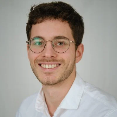 A young man with brown hair, glasses, and a light beard, smiling, wearing a white shirt against a neutral background.