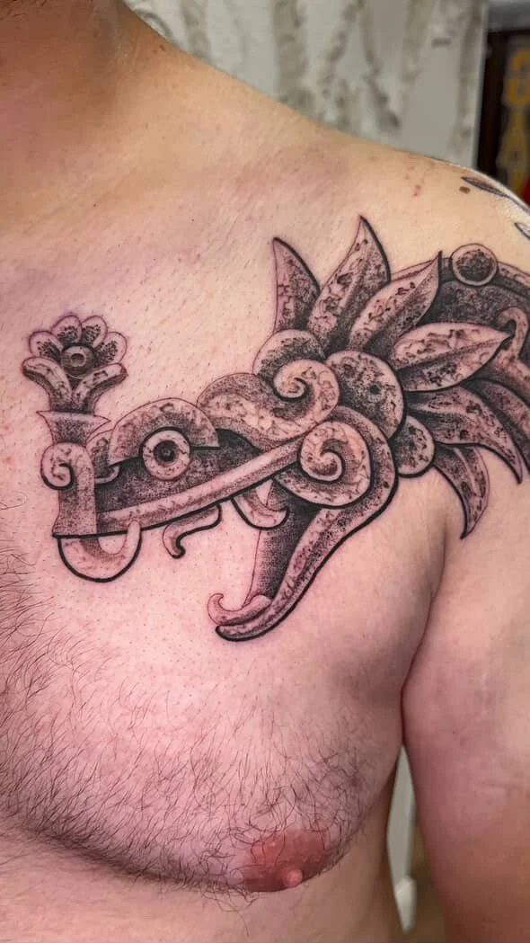 A black and gray tattoo of Quetzalcoatl, ornate design on a person's upper chest near the shoulder. The feathered serpent is tattooed in a unique way