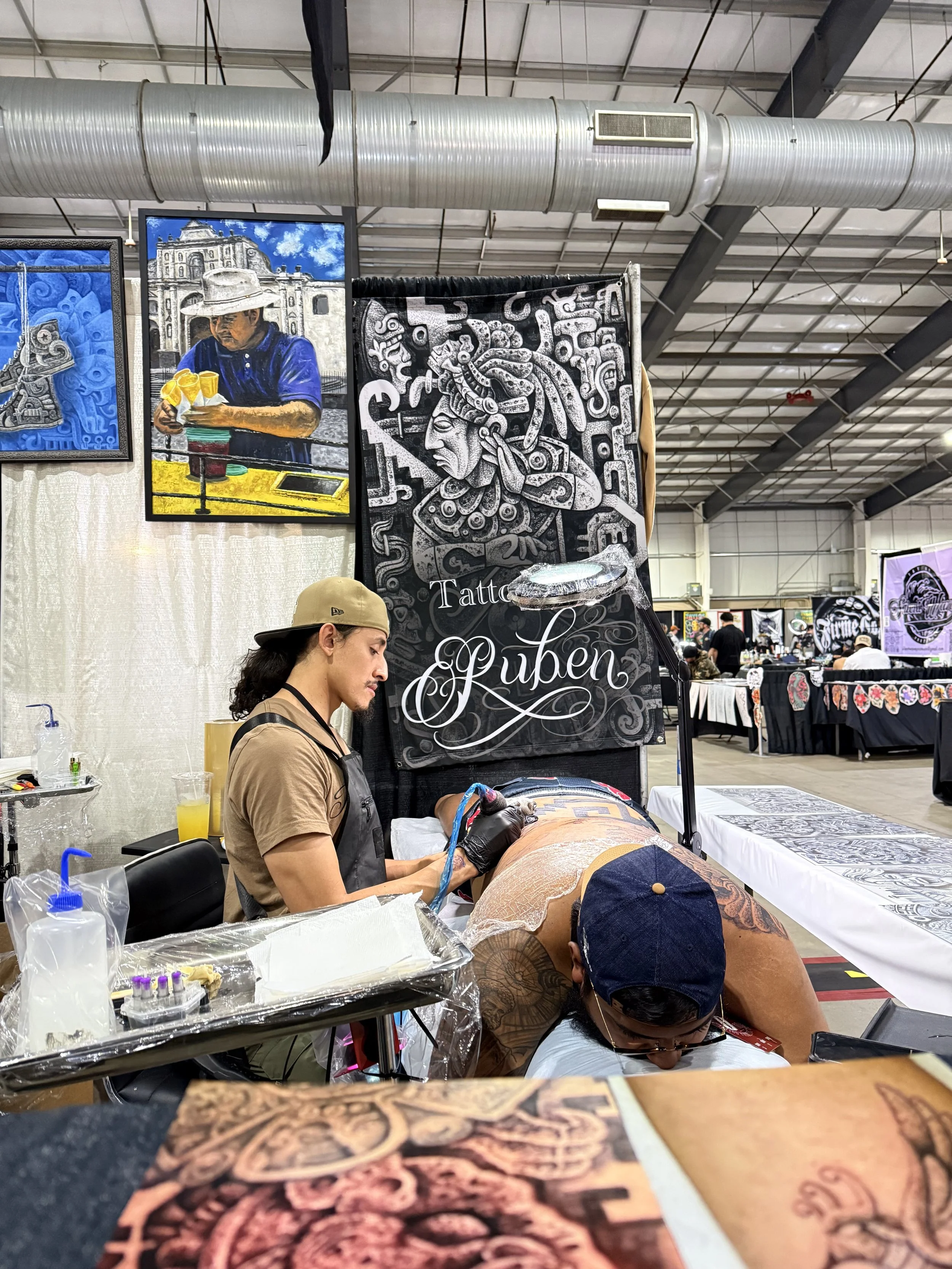 Tattoo artist Ruben Aguilar working on an Aztec back tattoo at an indoor tattoo convention in San Antonio, with artwork displayed on the walls behind. 