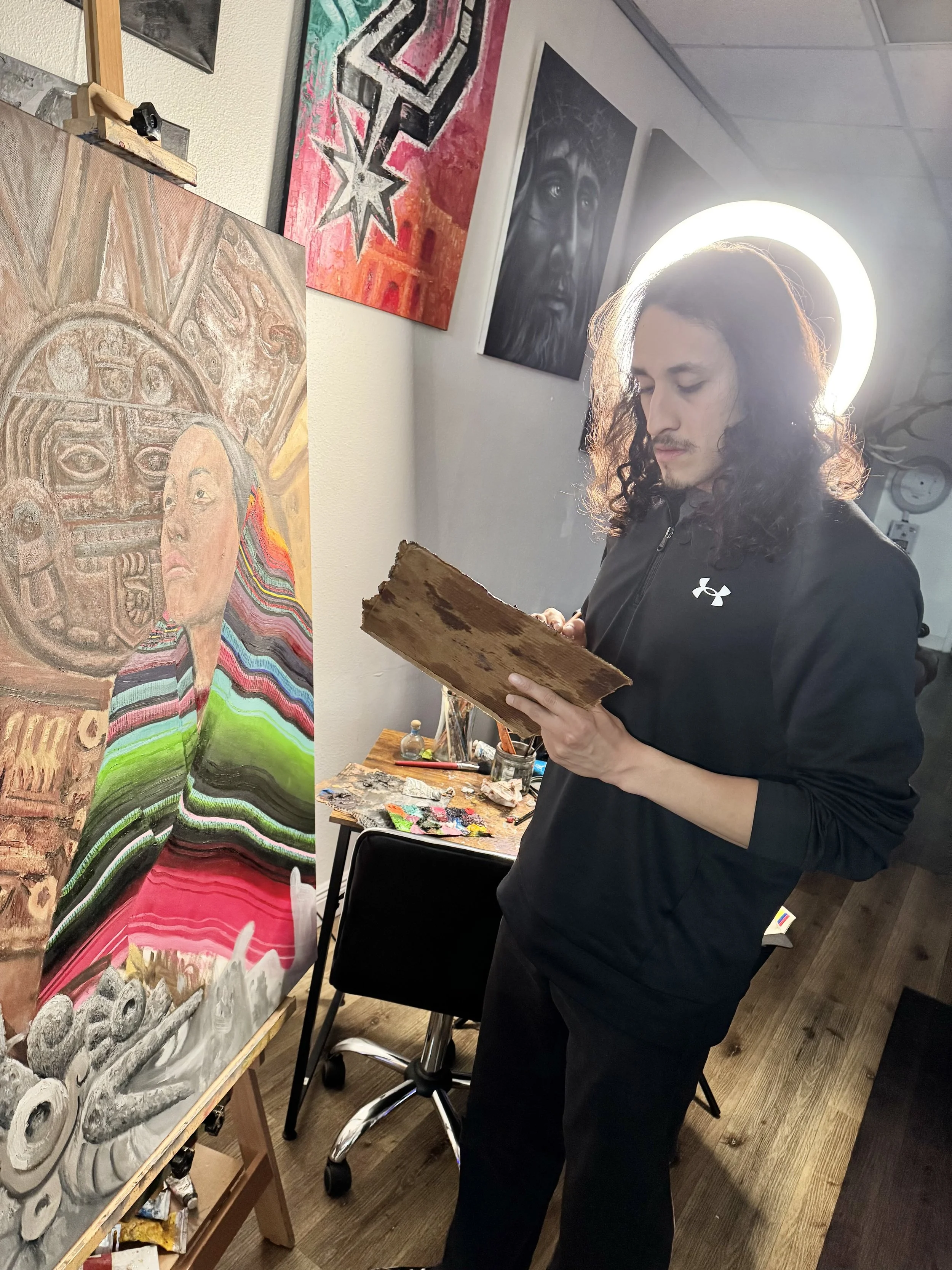 Ruben Aguilar stands in an art studio with multiple paintings on the walls. He is holding a wooden palette and appears to be contemplating his artwork, which is a colorful, abstract portrait of a woman on an easel. 