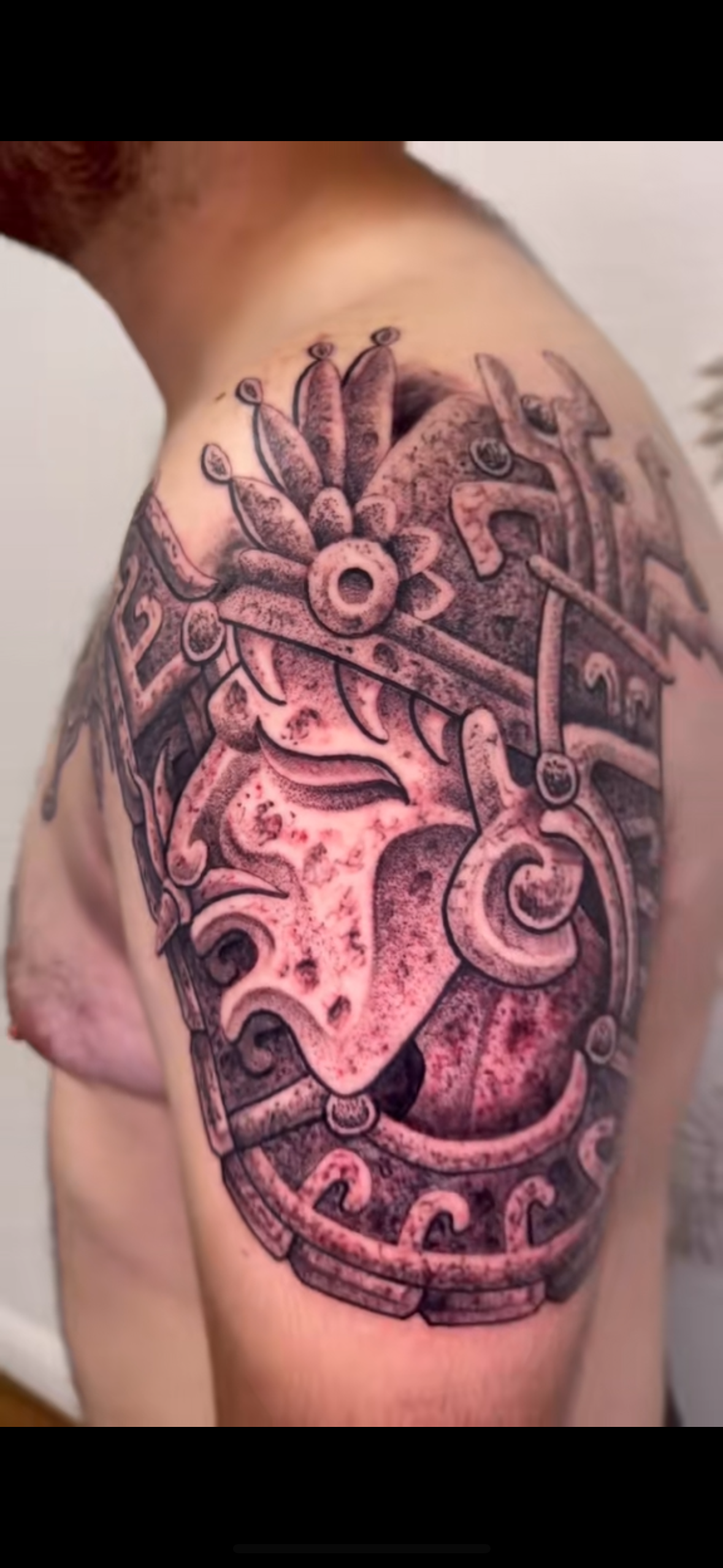 Close-up of a person's shoulder tattoo featuring intricate black and grey designs inspired by ancient or tribal patterns.