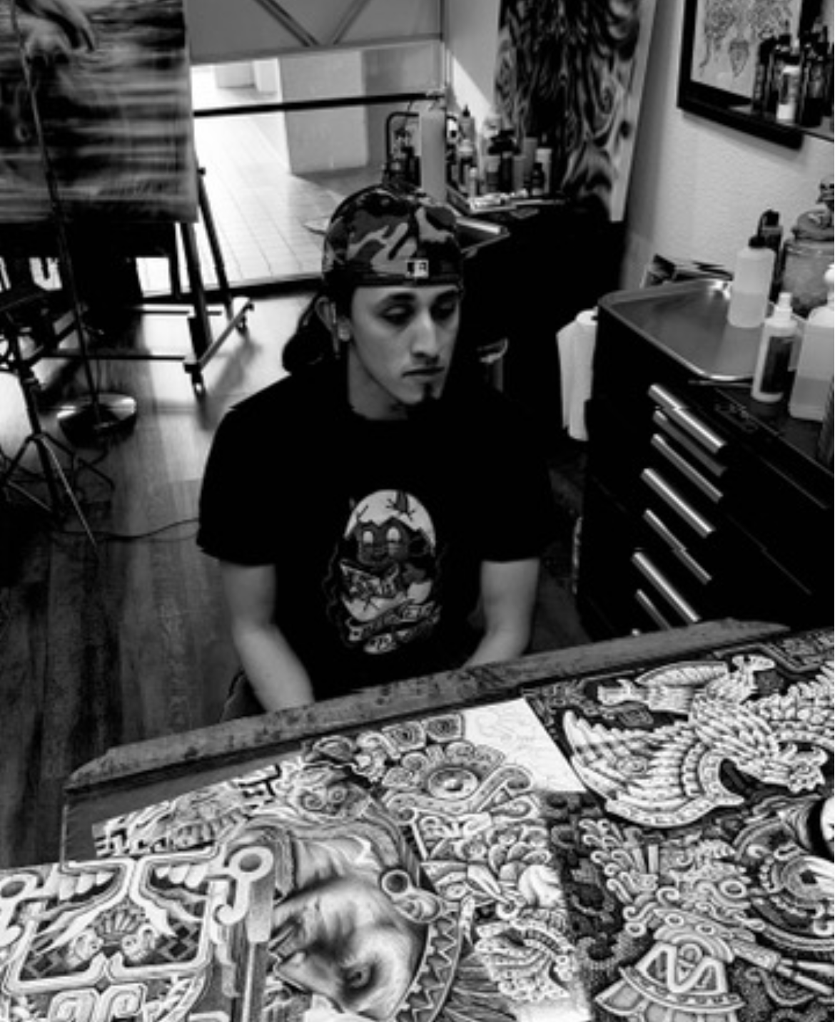 Tattoo artist Ruben Aguilar sitting with original hand drawn tattoo flash art, in a studio or art space with art supplies and artwork on the walls.