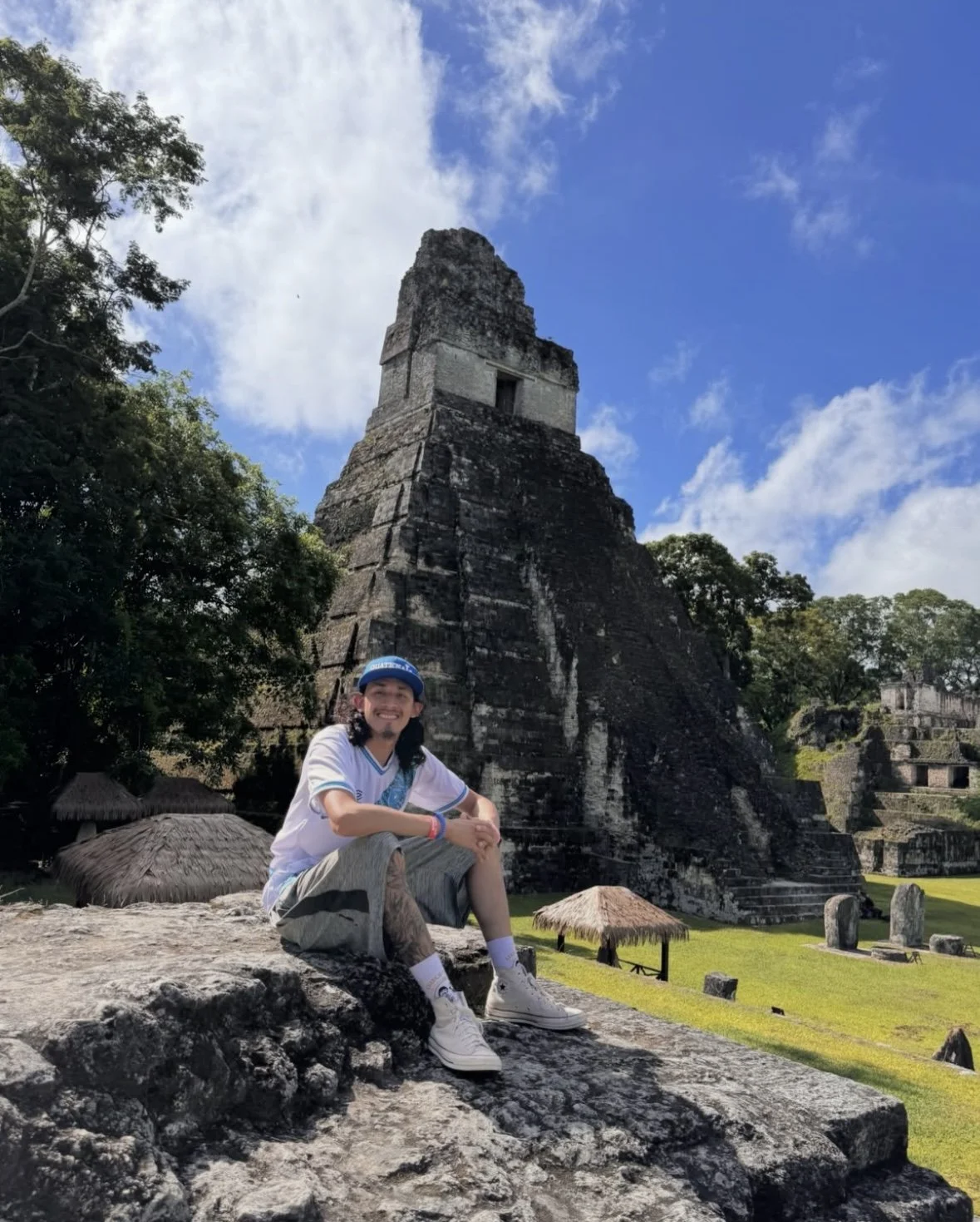 Ruben Aguilar sitting on the Mayan ruins with the ancient Mayan pyramids in Tikal behind him, in a lush green landscape under a partly cloudy sky.