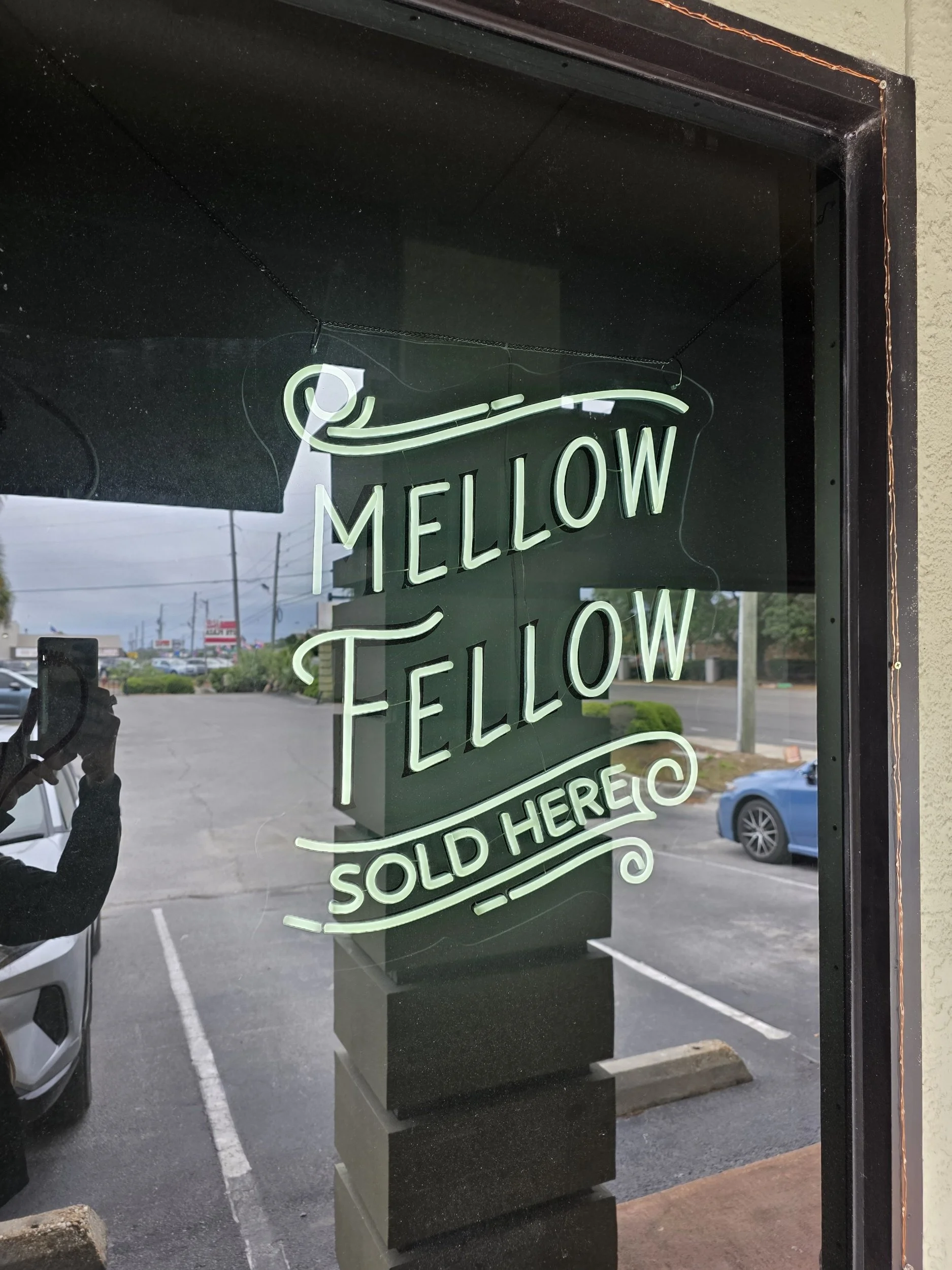 Neon sign on a window that reads 'MELLOW FELLOW SOLD HERE' with a wave design , reflecting on the glass.
