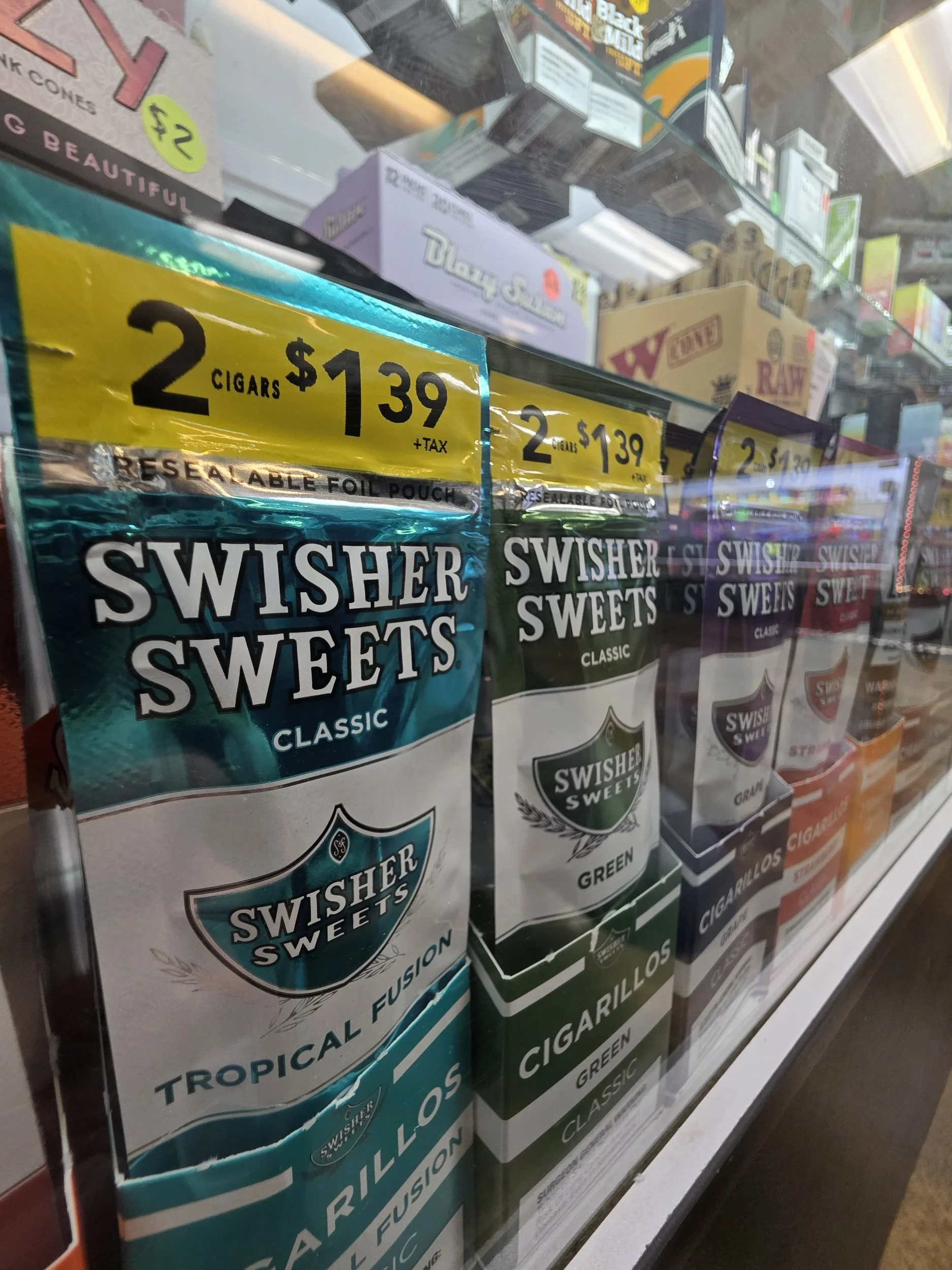 Display of Swisher Sweets cigar packages in a store window, showing various flavors like classic, green, and strawberry, with yellow promotional signs indicating a price of $1.39 for two cigars plus tax.