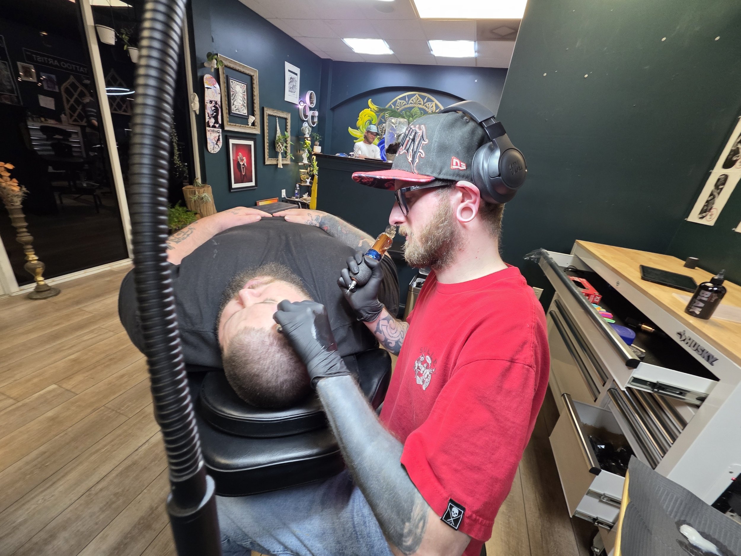 A man lying down on a chair while a tattoo artist applies a tattoo to the man's face. The background shows wall decor of framed pictures, plants, and shelves with decorative items.