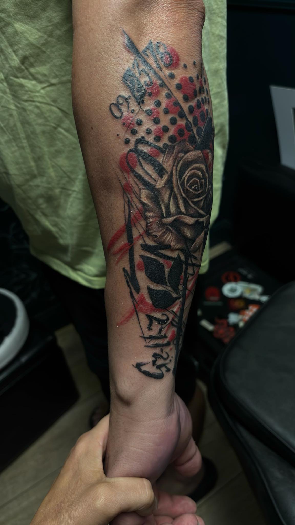 Trash polka style Tattoo of a rose with leaves, partly done in black and gray shading, with abstract red and black ink splashes and script writing above and below the flower.
