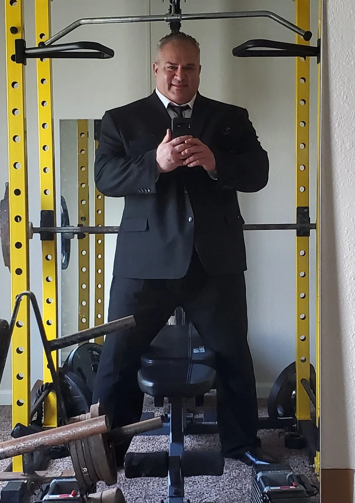 Man in a black suit and tie taking a selfie in a mirror, standing in a home gym with yellow and black workout equipment, weights, and a stool.