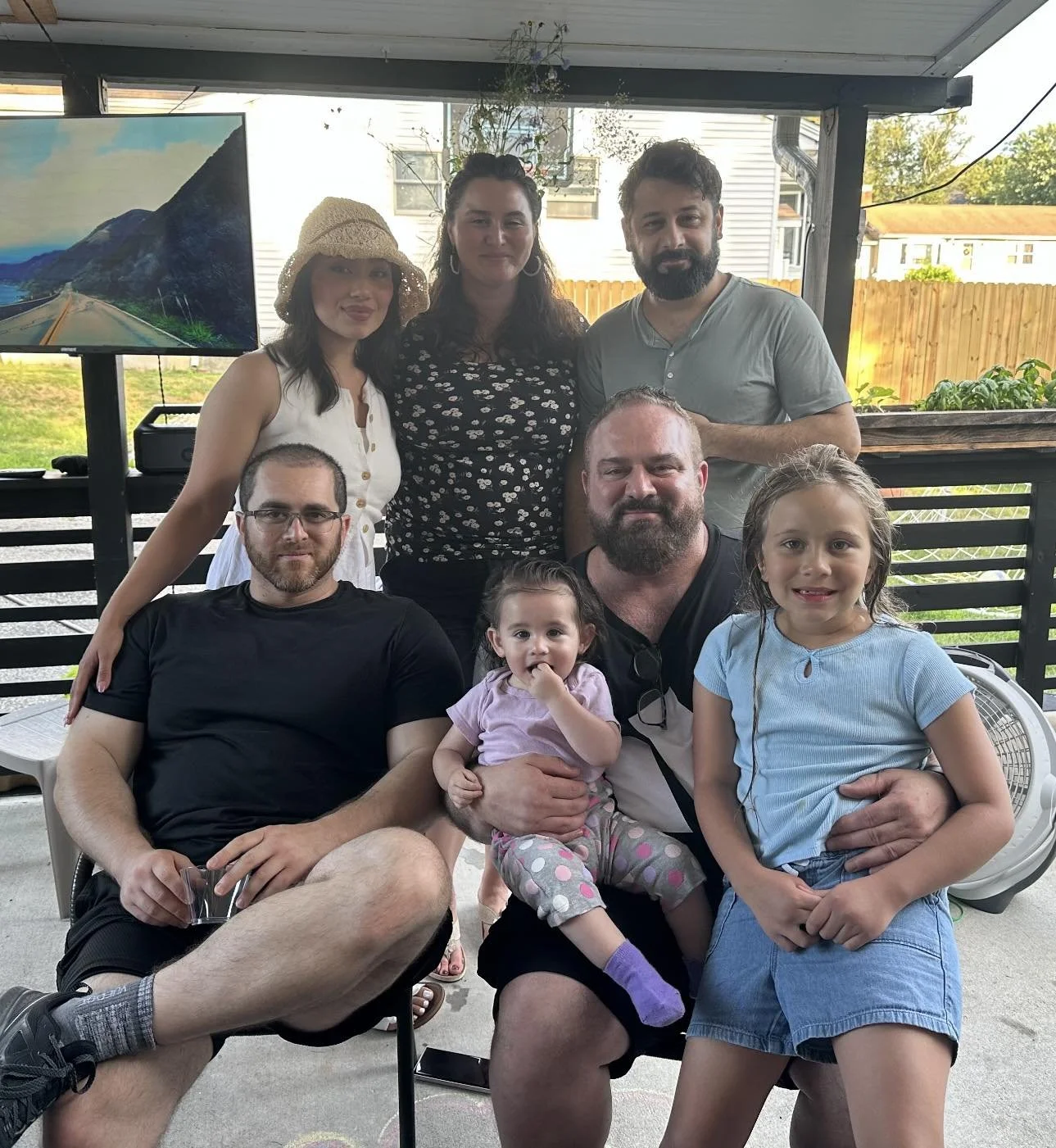 A group of seven people, including three children, gathered outside on a patio. They are sitting and standing, with a background of a wooden fence, some trees, and a house. There is a large screen on the left displaying a nature scene with mountains and sky.