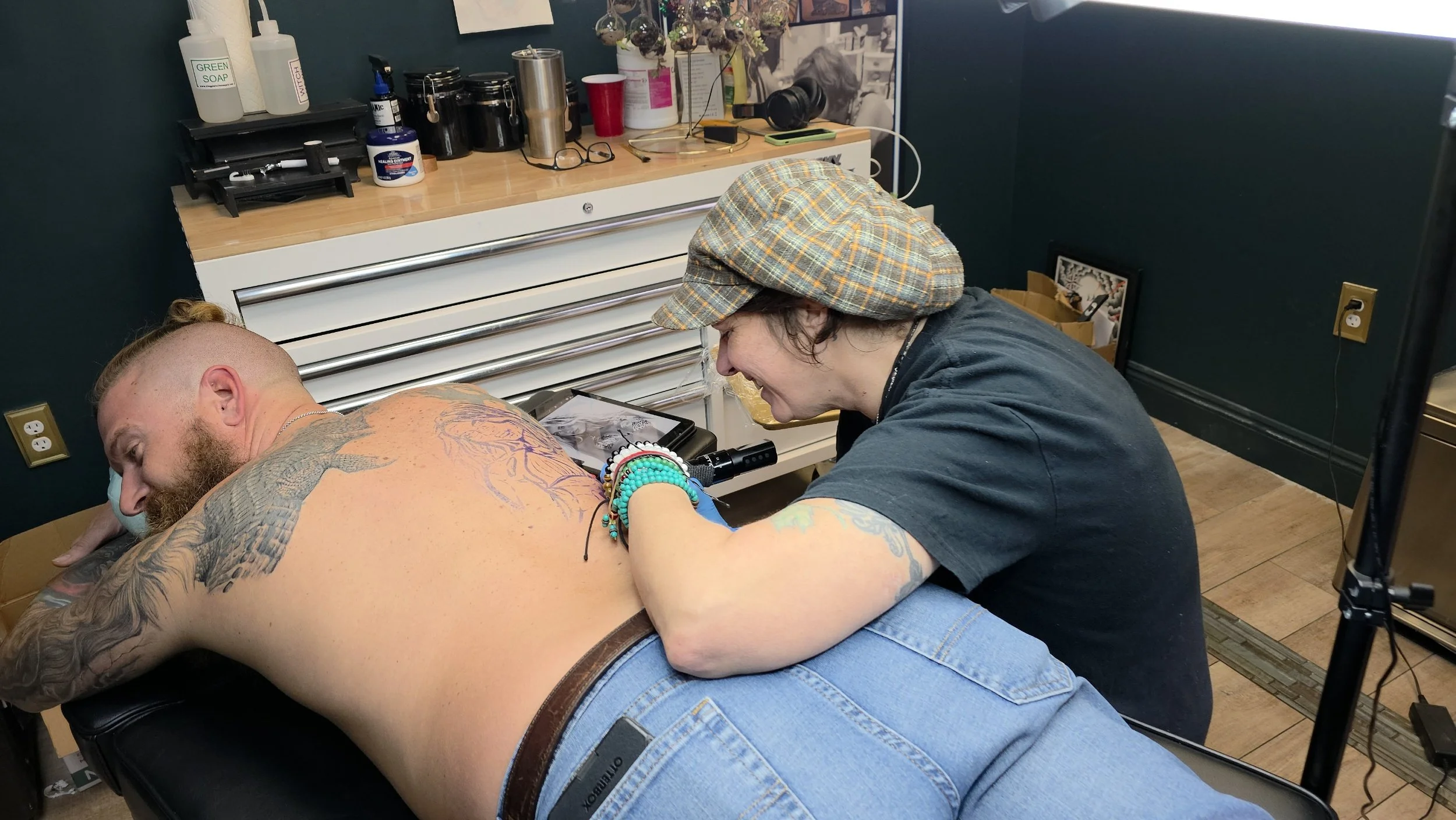 A person is getting a tattoo on their back by tattoo artist Crystal. The person is lying face down on a table, and the tattoo artist is focused on their work while smiling. The studio has various supplies and artwork on the walls.